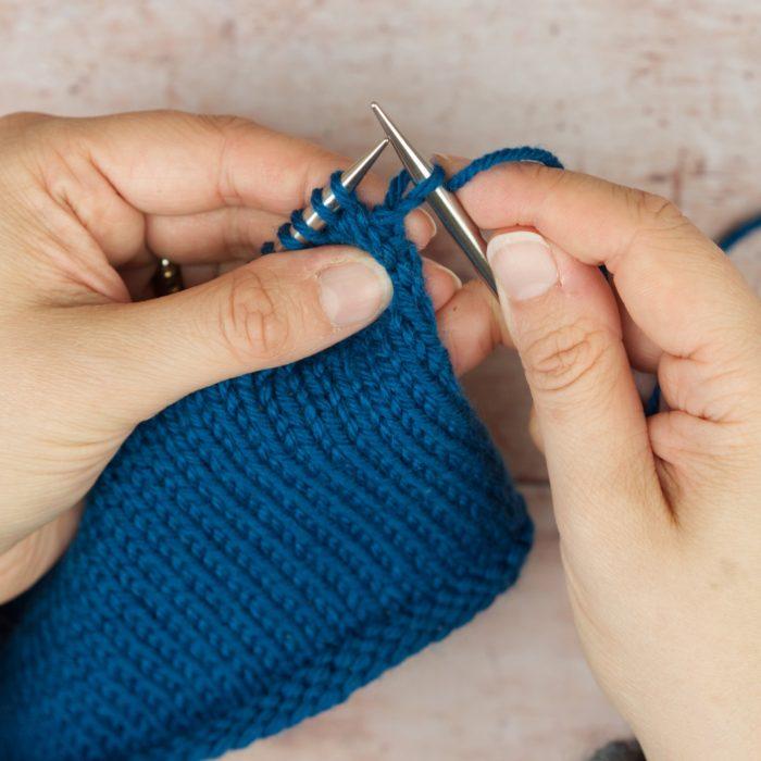 hands hold blue knitting and a knit stitch on right needle