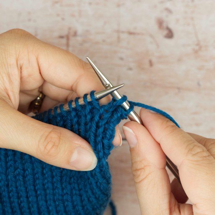 hands holding mid blue knitting with two stitches on right hand needle